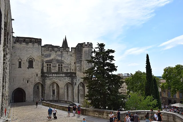 Los muros almenados y las torres de piedra del Palacio de los Papas vistos desde la plaza, con cipreses y visitantes
