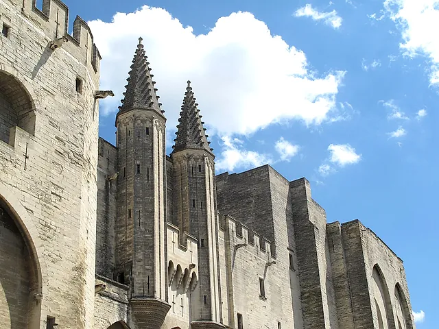 Due pinnacoli di pietra a punta e le mura merlate del Palais des Papes contro un cielo blu con nuvole bianche